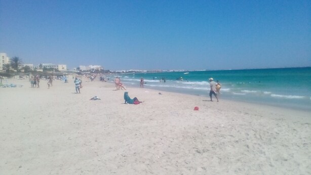 Ein Strand mit Menschen und Booten, Gebäude im Hintergrund und einem klaren Himmel.