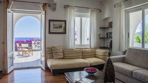 Un salon comprenant un canapé, une table basse et une bibliothèque. Il y a une grande fenêtre avec des rideaux, une porte donnant sur un espace extérieur avec une table et des chaises, et une vue sur la mer.