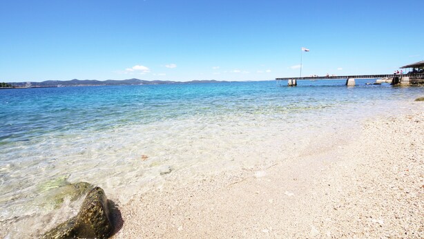 A beach with clear water, a sandy shore, and a pier with a flag. - PORAT Apartment-Breathtaking sunset view in Zadar, Croatia (Zadar)