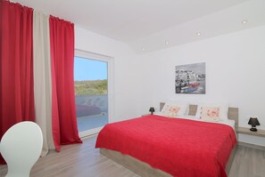 Ein Schlafzimmer mit einem großen Bett, einem Nachttisch, einem Sessel und Blick auf einen Balkon.