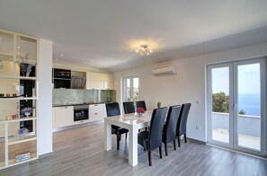 Eine moderne Küche mit weißen Schränken, ein Essbereich mit einem weißen Tisch und schwarzen Stühlen sowie ein Balkon mit Meerblick.