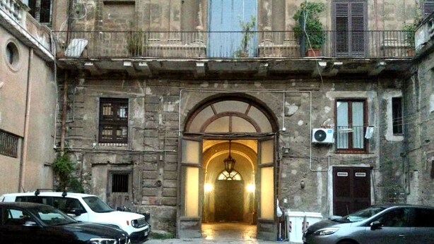 Ein Innenhof mit einem zentralen Bogen, der zu einem Gebäude führt, umgeben von parkenden Autos und Steinmauern.