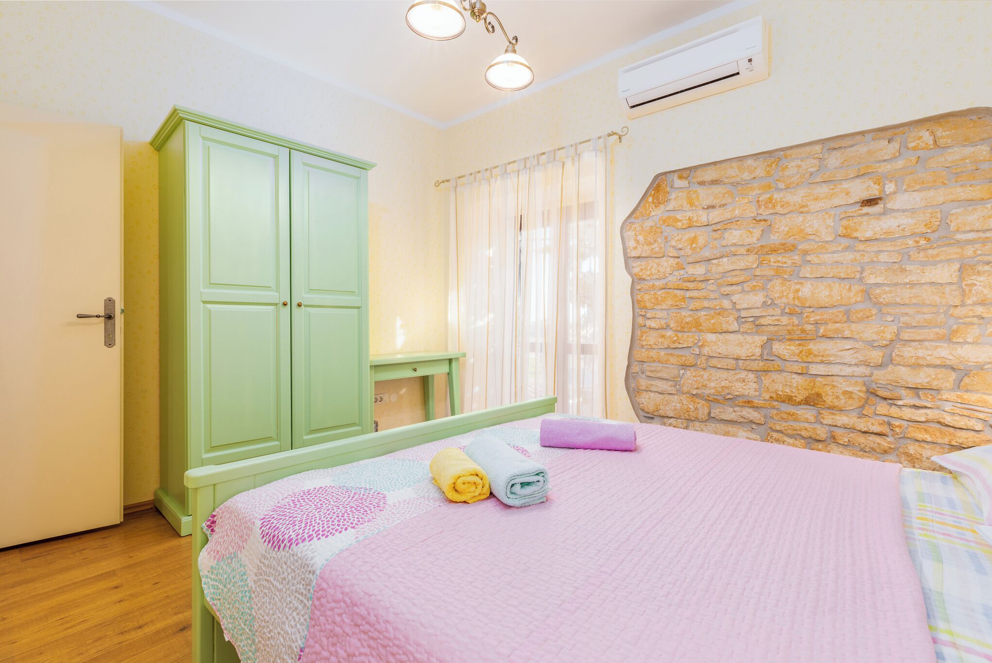 A bedroom with a bed, a green wardrobe, a stone wall, and a window with curtains.