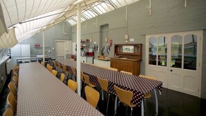 Zona pranzo con lunghi tavoli e sedie, angolo cucina con scaffali ed elettrodomestici, e vista attraverso una porta in vetro.