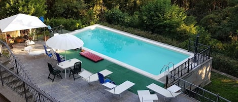 Una piscina rectangular con un deck verde, rodeada de abundante vegetación. Hay sillas de descanso blancas y una roja, junto con una mesa y sillas bajo un paraguas blanco.
