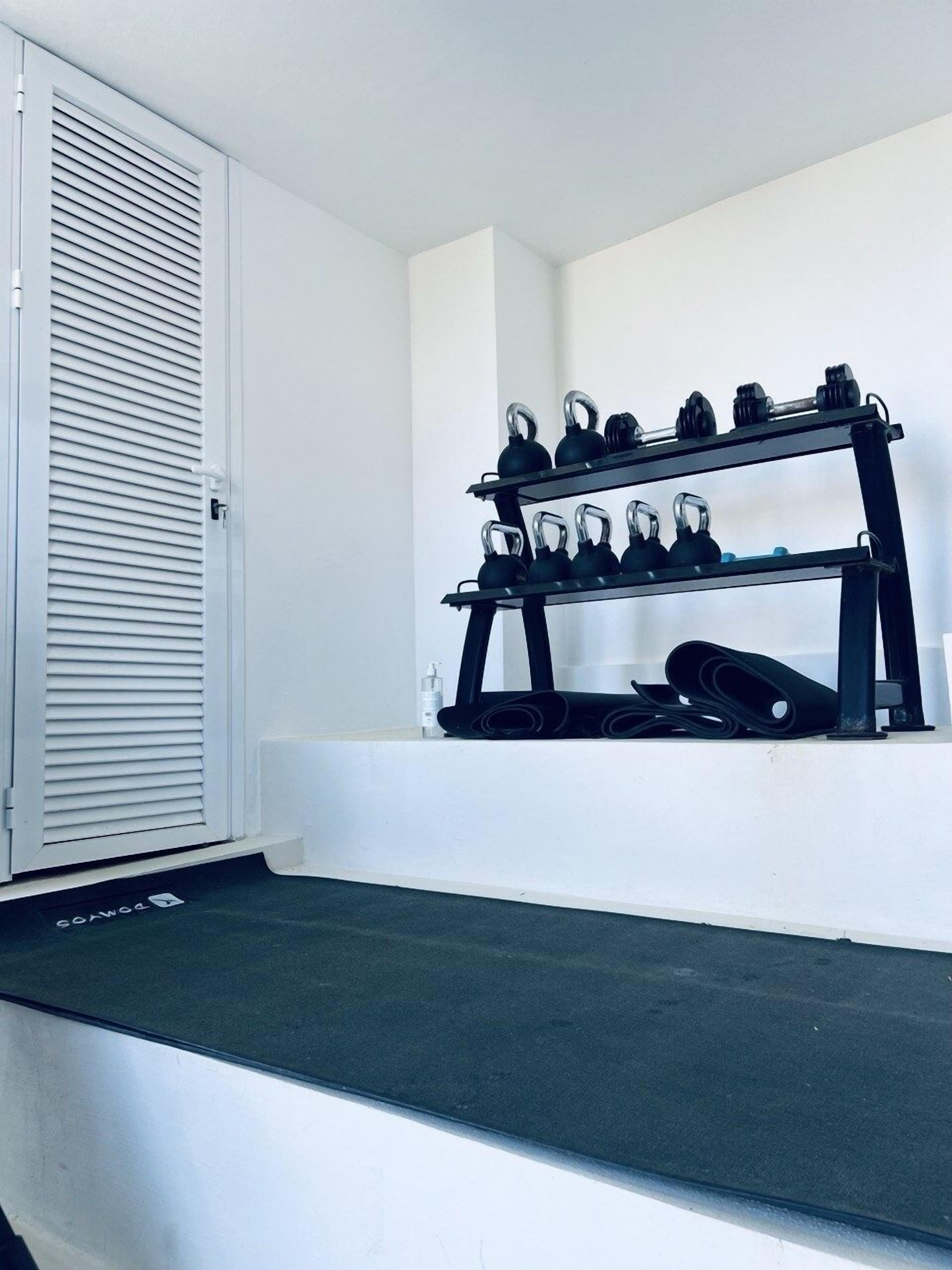 A room with a white wall, a black shoe rack with multiple pairs of shoes, and a white door with horizontal blinds.