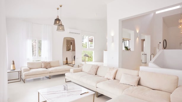 Een ruime woonkamer met een grote witte bank, een salontafel en een eethoek. Twee hanglampen zijn aan het plafond bevestigd.