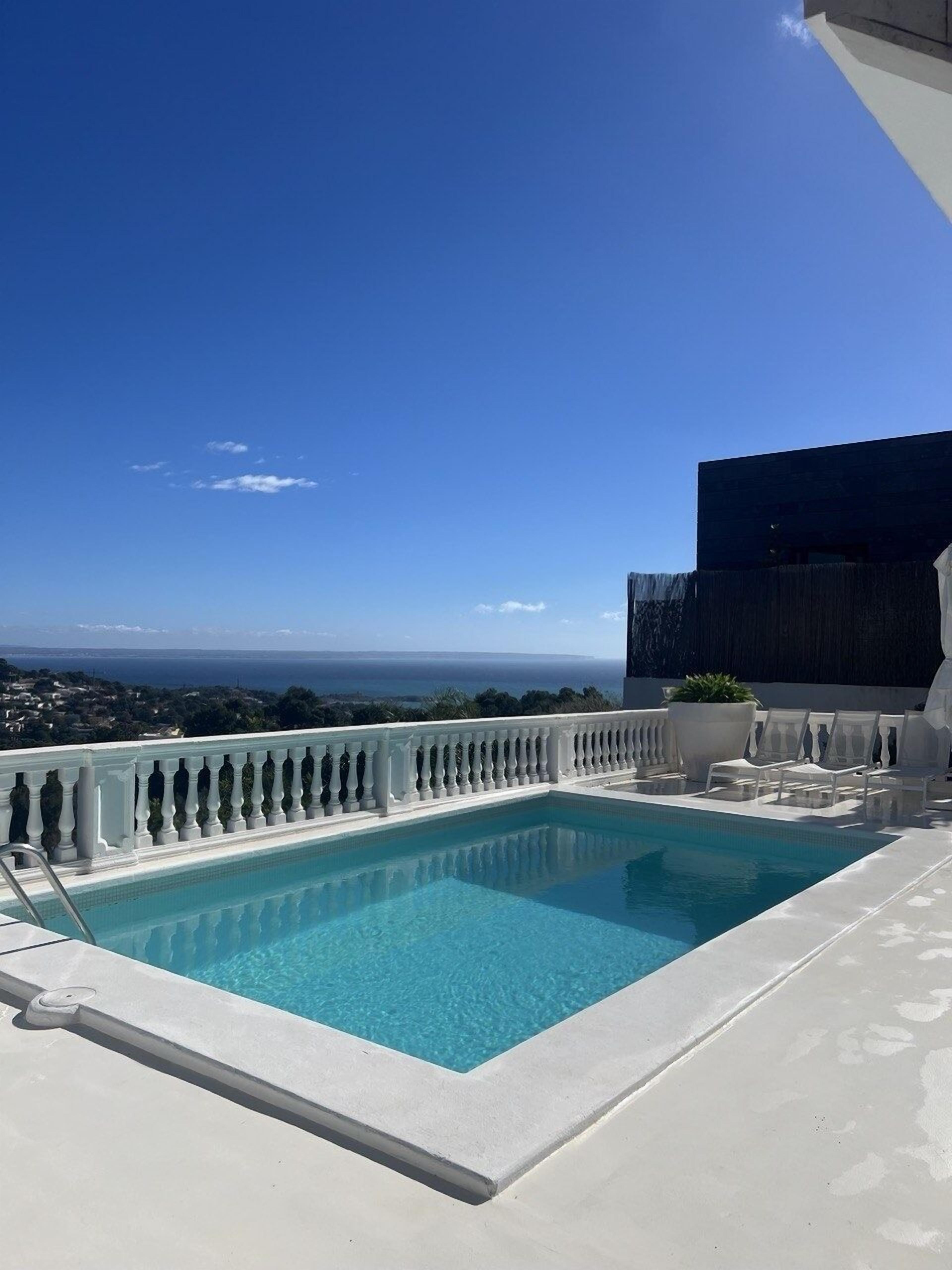 A rectangular pool with a white balustrade, surrounded by white lounge chairs and a potted plant, overlooking a coastal view.