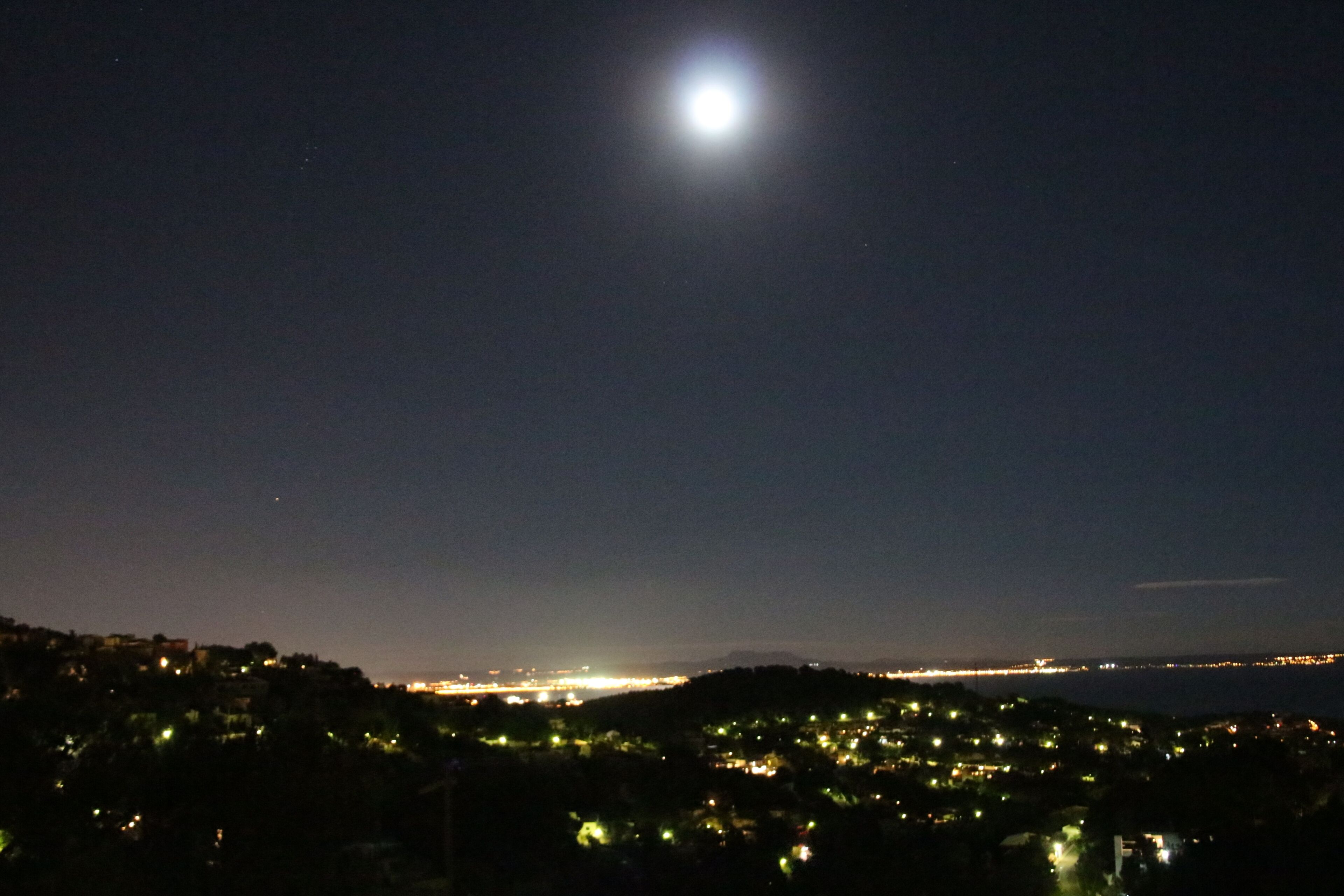 Vista nocturna de un pueblo costero, con una luna grande en el cielo.