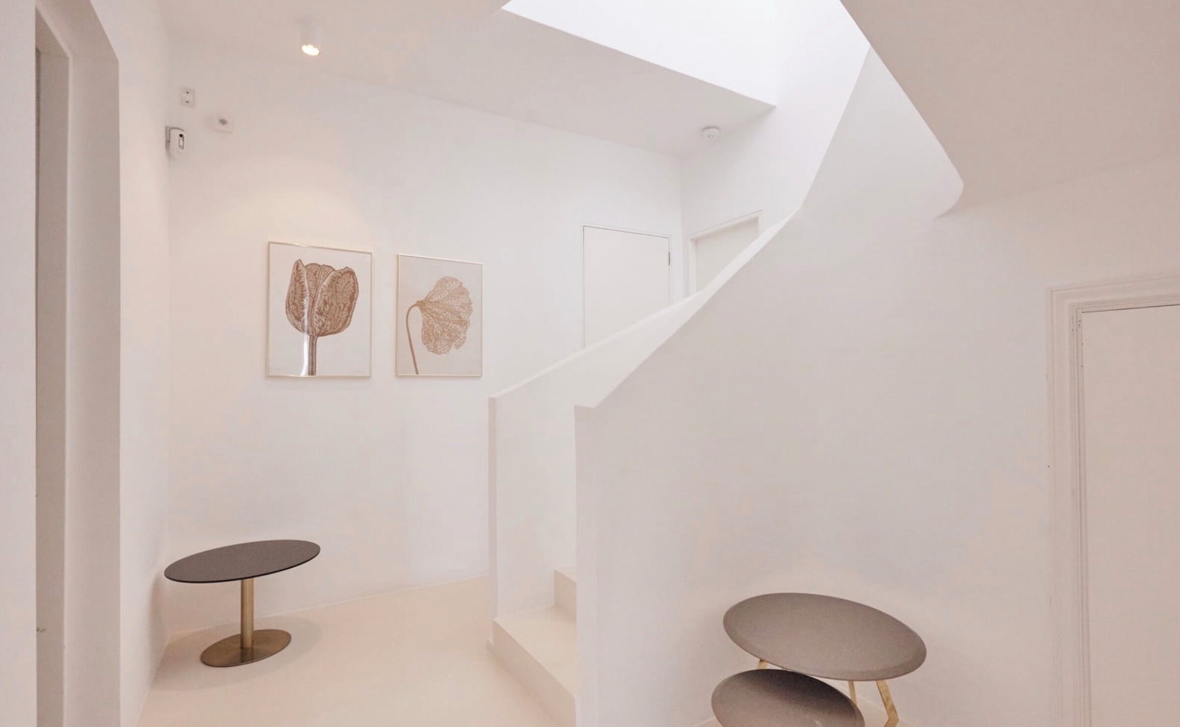 Un intérieur moderne avec un escalier menant à un deuxième étage, une décoration minimaliste et deux reproductions botaniques encadrées accrochées au mur.