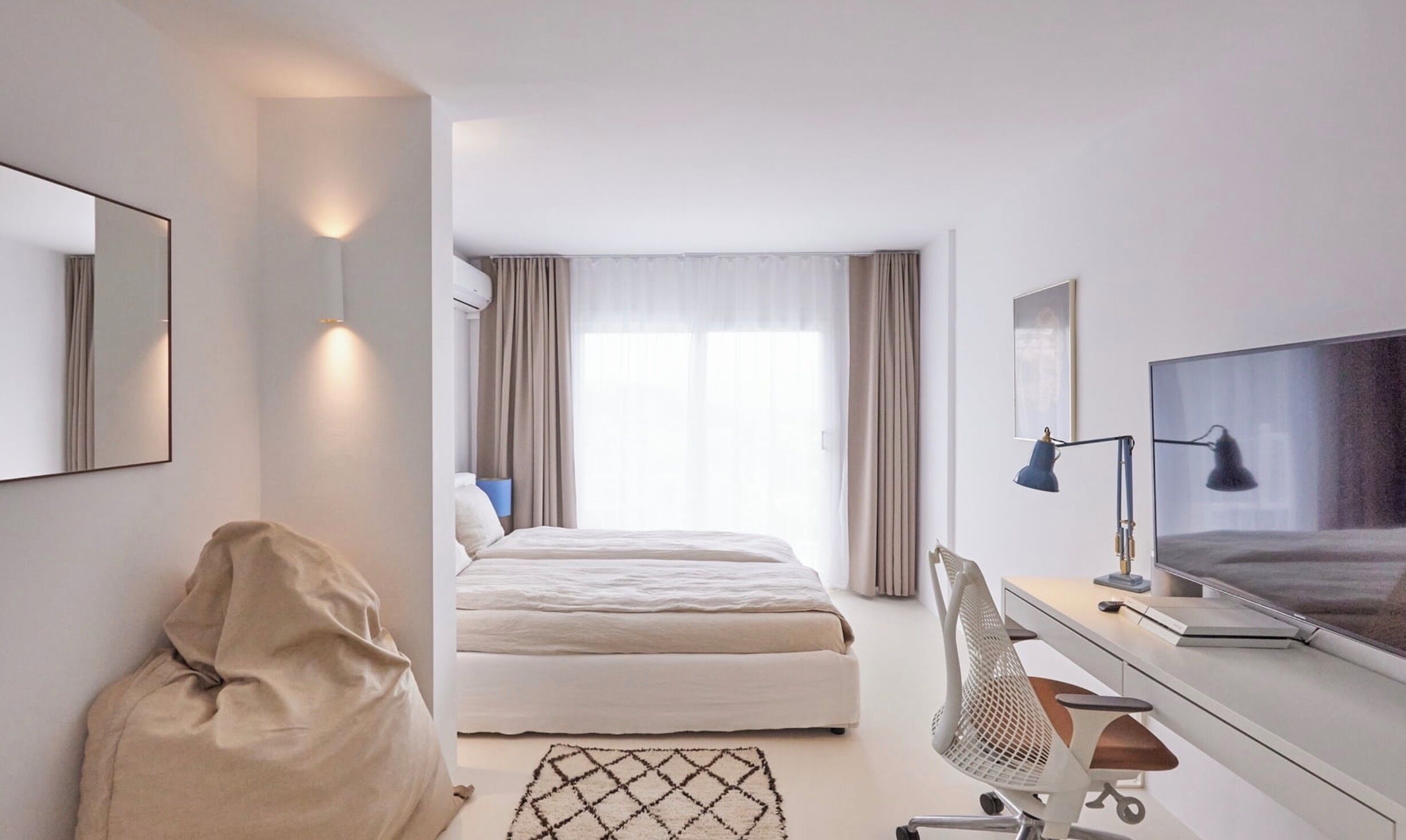 A modern hotel room with a bed, a desk with a computer, a TV, and a large window with curtains.