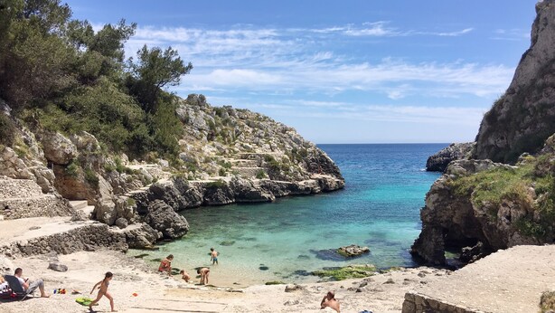 Uma praia rochosa com água cristalina, pessoas tomando sol e um caminho que leva ao mar.