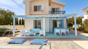 A two-story house with a covered patio, white furniture, and a pool area. - This amazing luxury villa is located in the heart of Protaras just 4 minutes walk to the Main Strip (Protaras)