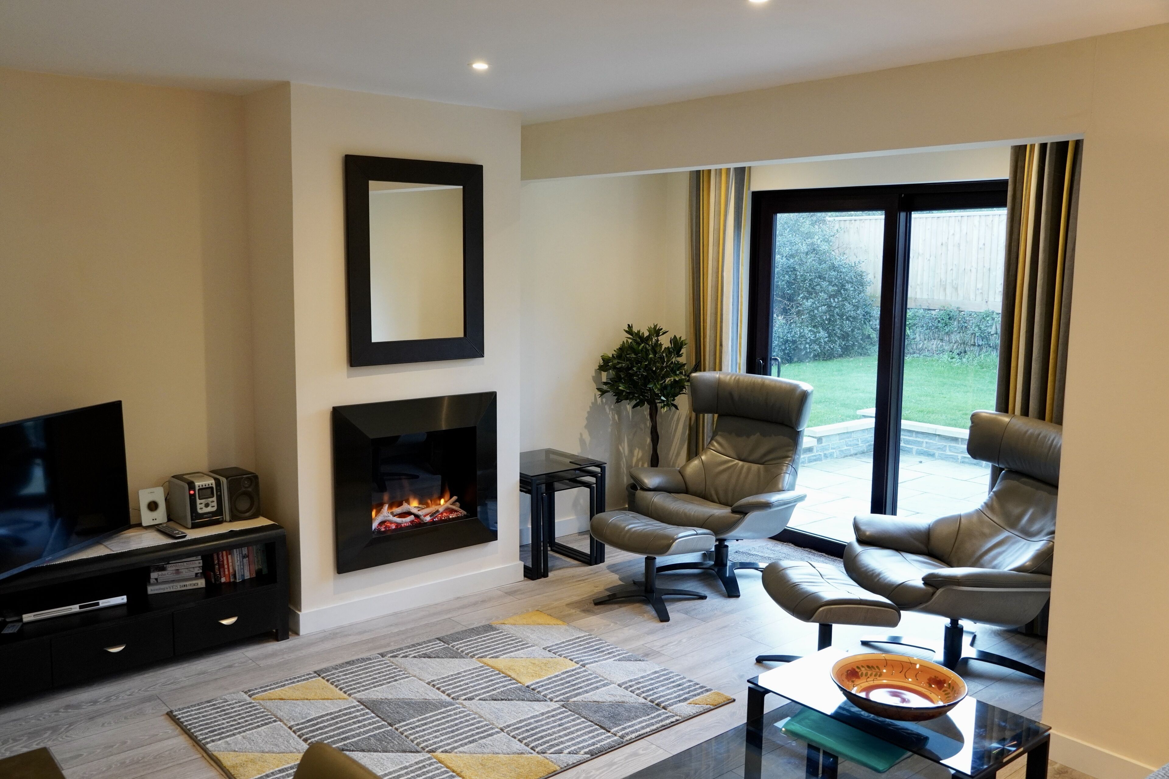 A modern living room with a flat-screen TV, a fireplace, leather armchairs, and a large window with curtains.