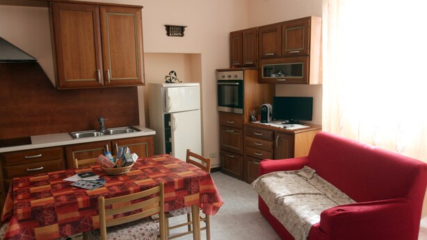 Cocina compacta con armarios de madera, un refrigerador, un horno y un microondas. Hay una mesa de comedor con sillas y un sofá rojo en la sala de estar.