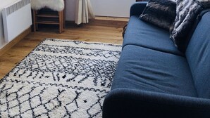 Un salon chaleureux avec un canapé bleu, un fauteuil blanc à poils longs, un tapis à motifs et une grande fenêtre avec des rideaux.