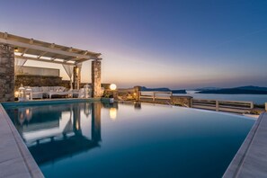 A serene infinity pool with a stone deck, outdoor seating, and a view of the sea at dusk. - 5 bedroom luxury villa with pool and stunning sea views and amazing sunsets (Akrotiri Thiras)