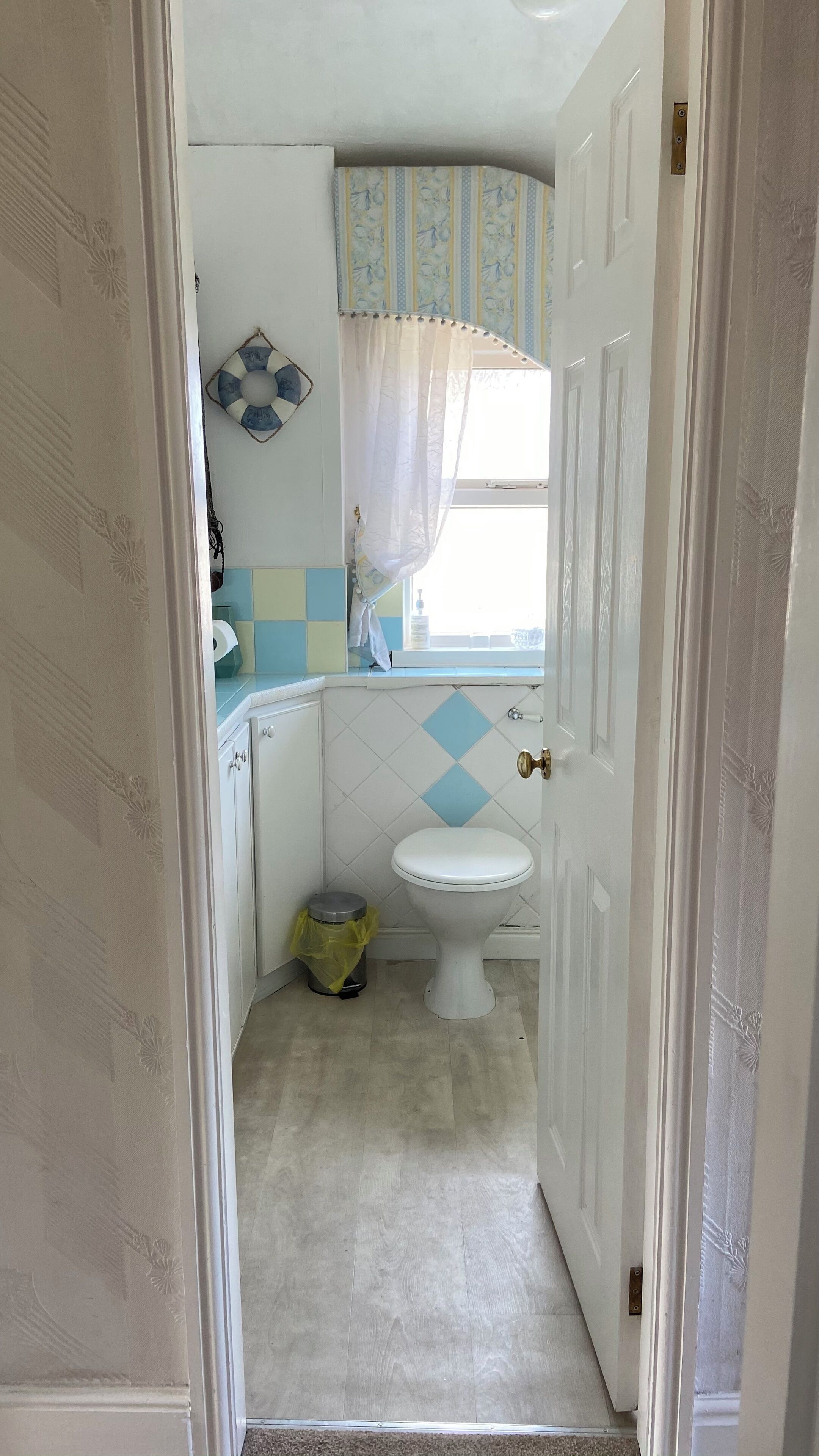 A bathroom with a toilet, a trash can, and a window with a curtain.