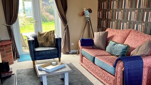 Una sala de estar con un sillón de cuero, un sofá con cojines estampados, una mesa de centro con una bandeja, una estantería y una ventana con cortinas.
