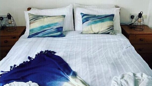 Una habitación de hotel con una cama, dos mesitas de noche, un cuadro de un faro y una manta azul.