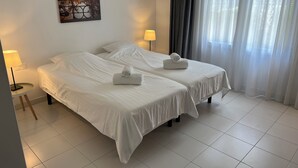 A hotel room with two beds, a nightstand, a lamp, a painting, and a window with curtains.