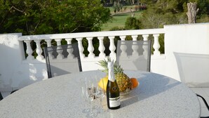 A table with a bottle of champagne, glasses, a pineapple, and a bowl of fruit on a balcony overlooking a golf course.