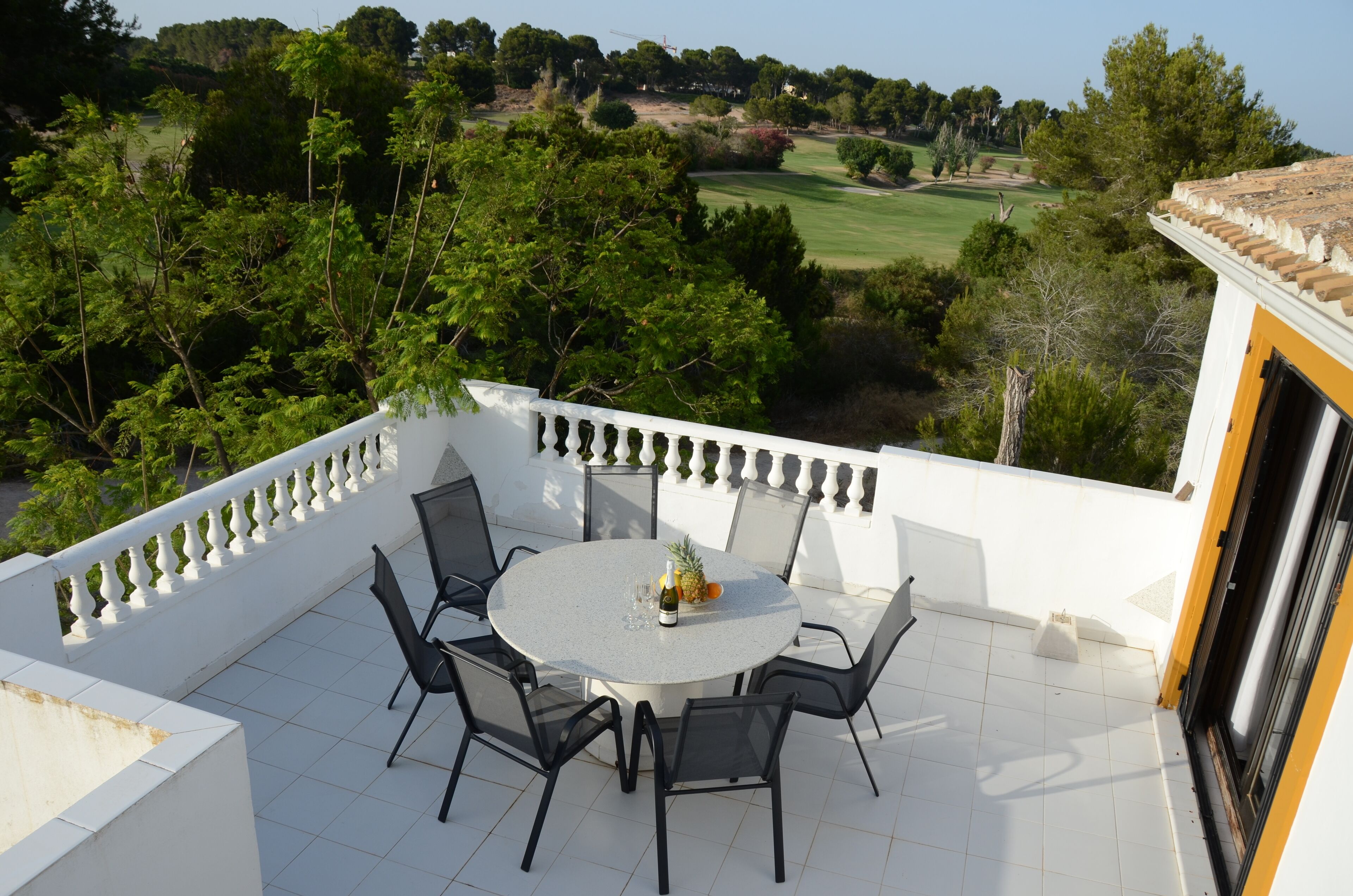 Een ronde tafel met stoelen op een balkon met een witte reling, met uitzicht op een golfbaan en groen.