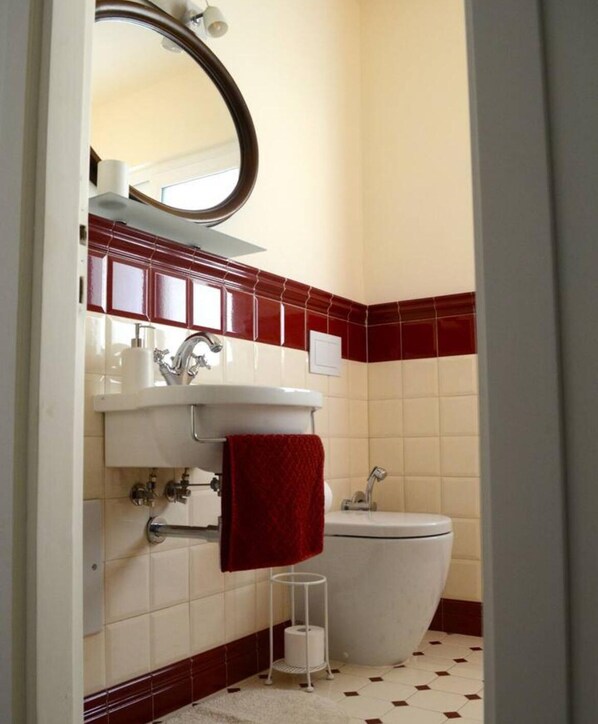 Ein Badezimmer mit einem weißen Waschbecken, einem Spiegel, einem roten Handtuch, einer Toilette und einem kleinen weißen Hocker.