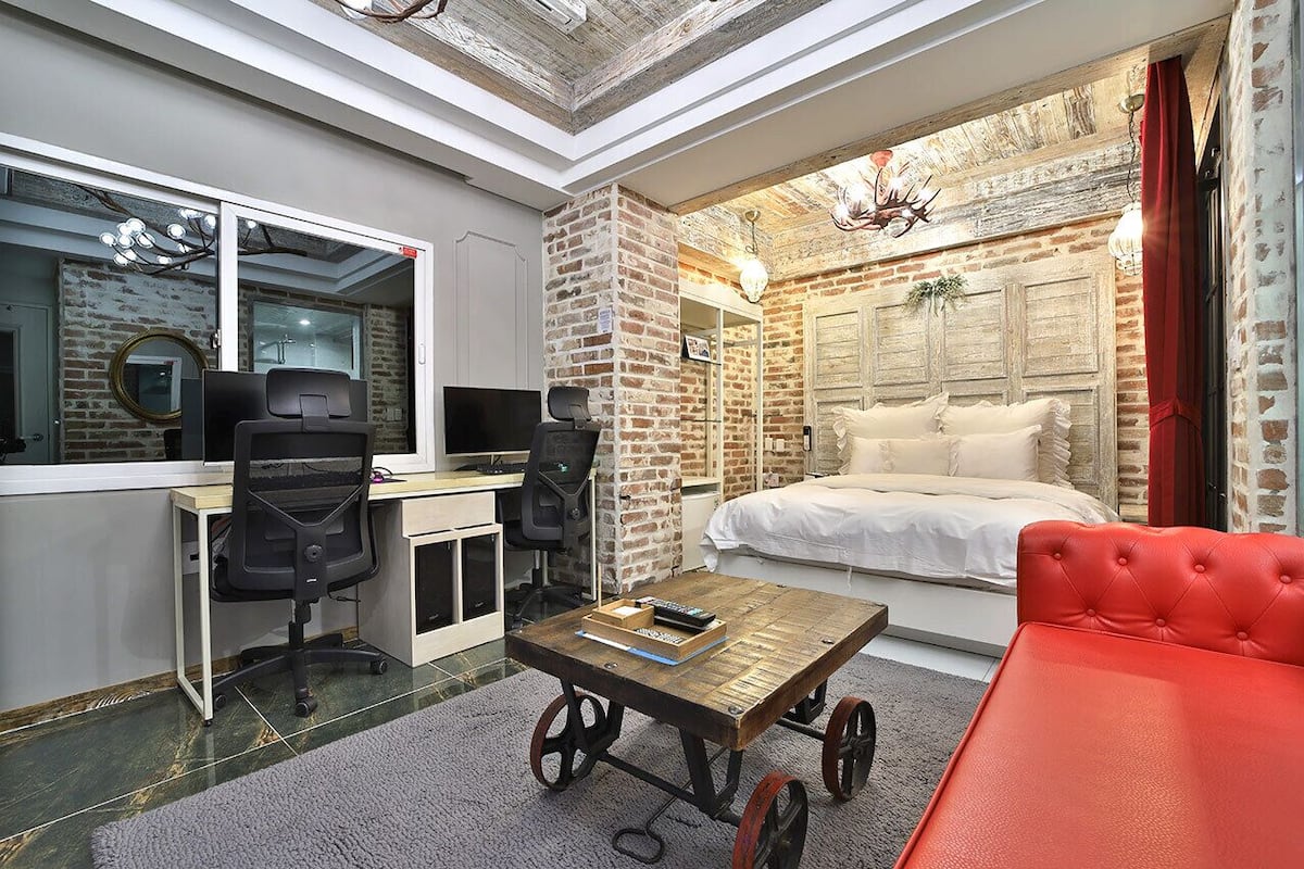 Un dormitorio moderno con pared de ladrillo, una cama, un escritorio con ordenador y un sofá rojo.