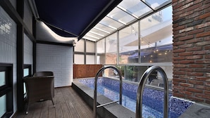 Un espacio exterior cubierto con vidrio, una terraza de madera, una piscina pequeña y una silla de mimbre.