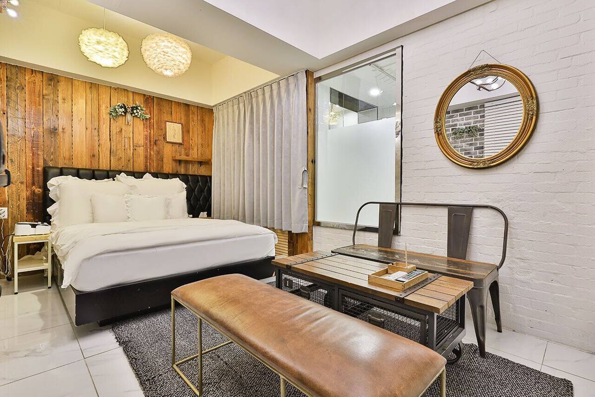 Un dormitorio moderno con cabecera de madera, una cama grande, una mesita de noche, un banco y un espejo en la pared.