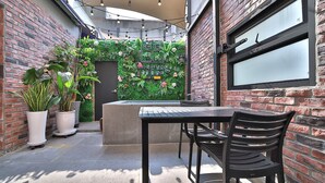 Un patio con pared de ladrillo, plantas en macetas, una mesa con sillas y una ventana.
