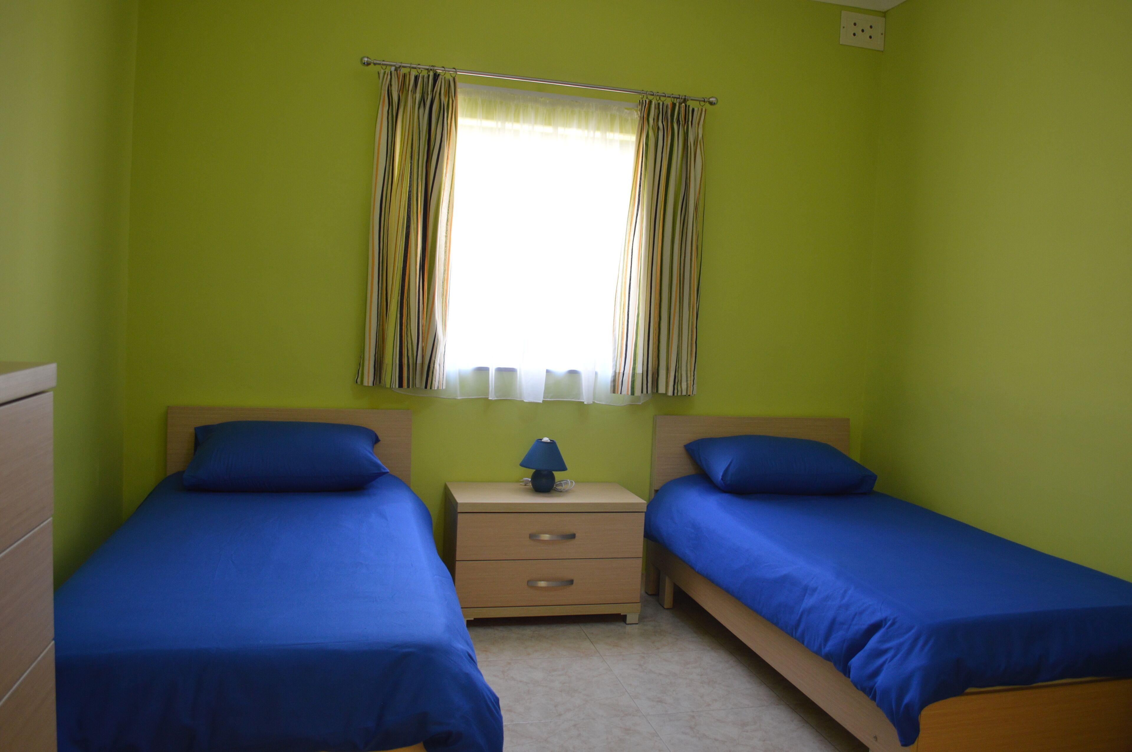 A room with two single beds, a nightstand with a lamp, and a window with curtains.