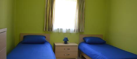 A room with two single beds, a nightstand with a lamp, and a window with curtains.