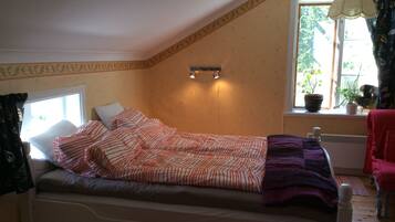 A single bed in a room with a sloped ceiling, a window with curtains, and a potted plant on the windowsill.