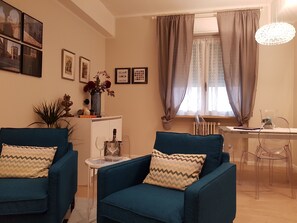Un salon avec deux fauteuils bleus, une petite table sur laquelle se trouvent une bouteille et des verres, un vase rempli de fleurs et un coin repas avec une table et des chaises.