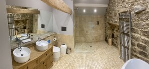 A bathroom with a stone wall, a wooden vanity, two sinks, a shower, and a metal towel rack.