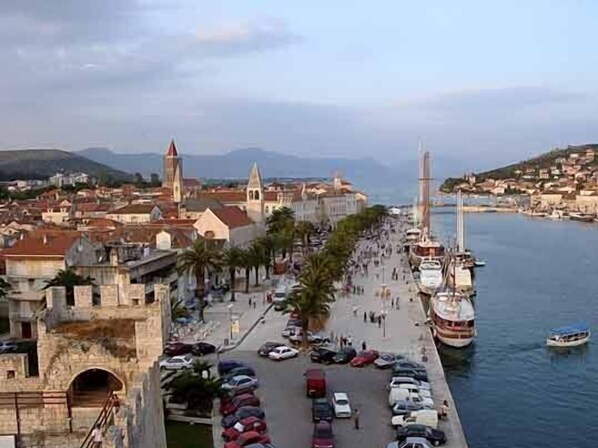 A waterfront promenade with parked cars, a large boat docked on the right, and historic buildings with towers in the background.