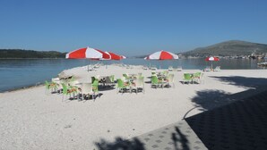 Ein Café direkt am Strand mit Außenbestuhlung und Sonnenschirmen, mit Blick auf ruhiges Gewässer.