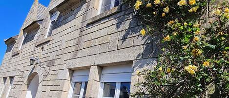 A stone building with a steeply pitched roof, multiple windows, and a flowering bush.