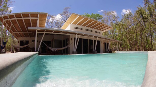 Un'area piscina moderna con una piscina blu cristallina, una zona relax coperta e vista sugli alberi circostanti.