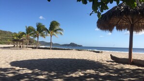 Una spiaggia con palme, una costruzione a tetto di paglia e vista sull'oceano e su un'isola in lontananza.