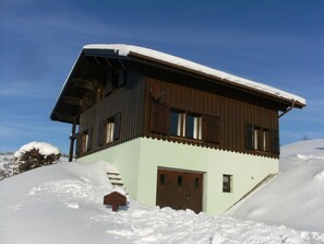 A two-story wooden cabin with a sloped roof covered in snow, a wooden door, and a small window.