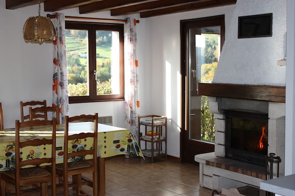 A dining area with wooden chairs, a table with a fruit-themed tablecloth, a bookshelf, and a fireplace with a visible fire.
