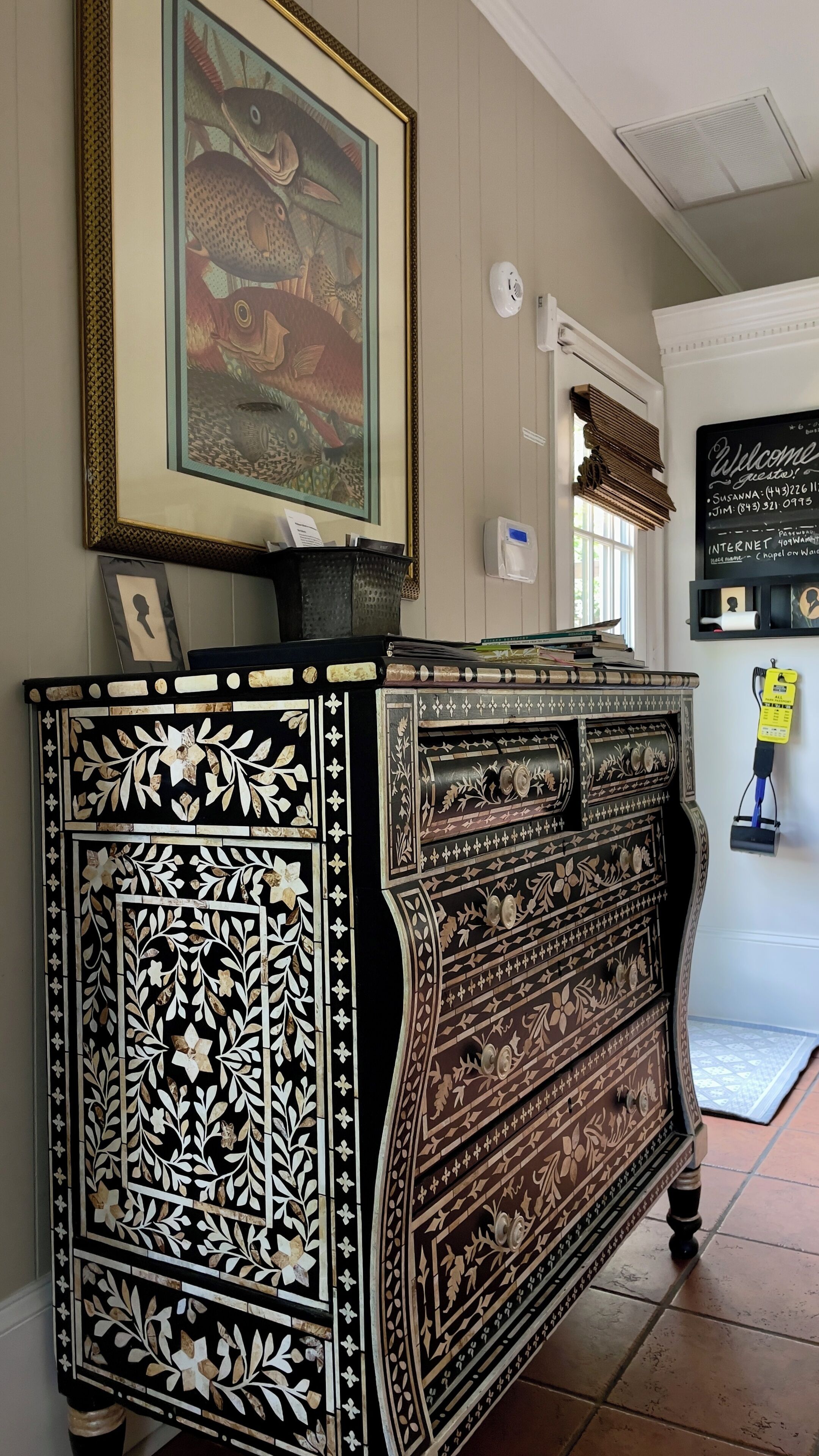 A black and white ornate cabinet with intricate floral patterns, a framed fish painting above it, and a chalkboard with a welcome message.