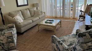 Ett vardagsrum med en beige soffa, två fåtöljer, ett soffbord i glas och utsikt över havet genom tunna, genomskinliga gardiner.