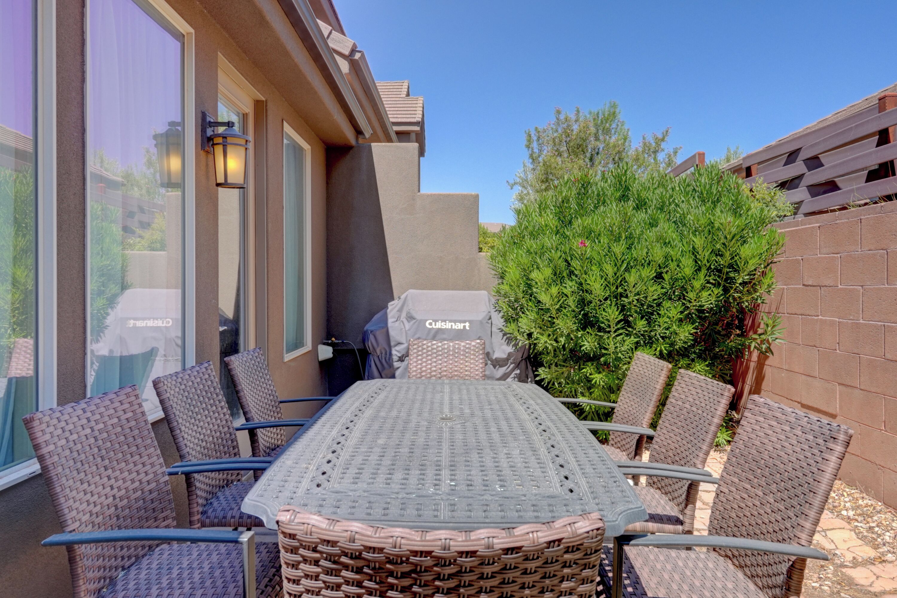 An outdoor dining area with a table and chairs, a grill, and a view of a building with large windows and a brick wall.