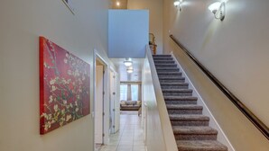 Un pasillo con una escalera alfombrada, un cuadro floral rojo en la pared y vistas a una sala de estar con sofá y ventana.