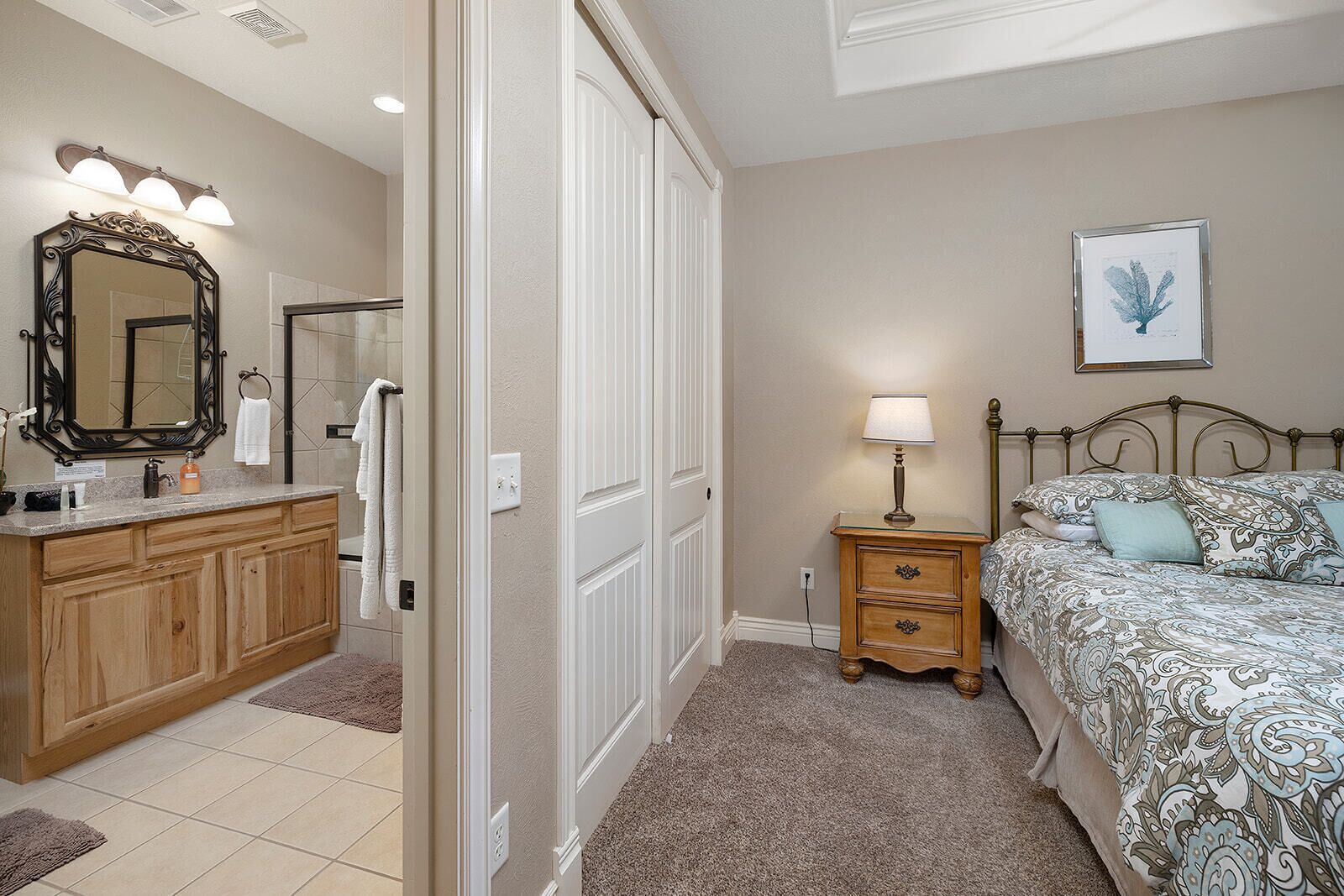A bedroom with a bed, bedside table, and a nightstand. There is a bathroom with a vanity, mirror, and towel rack.