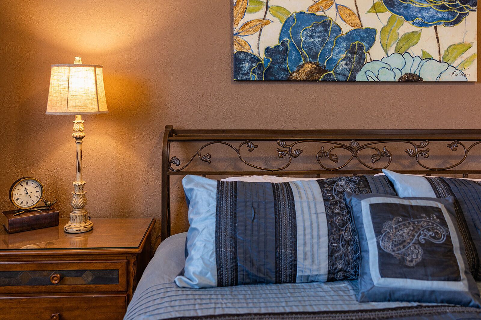 A neatly made bed with multiple pillows, a wall clock, a lamp, and a floral painting on the wall.
